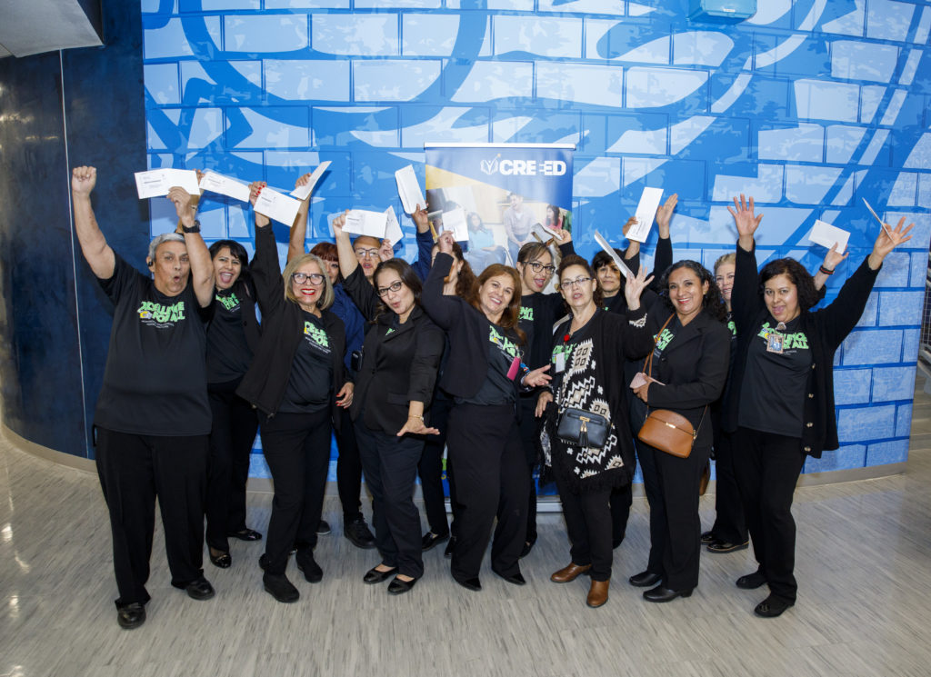 A group of people from Montwood middle school smiling in front of a CREEED pop up banner holding their white envelopes containing their CREEED gift cards in the air