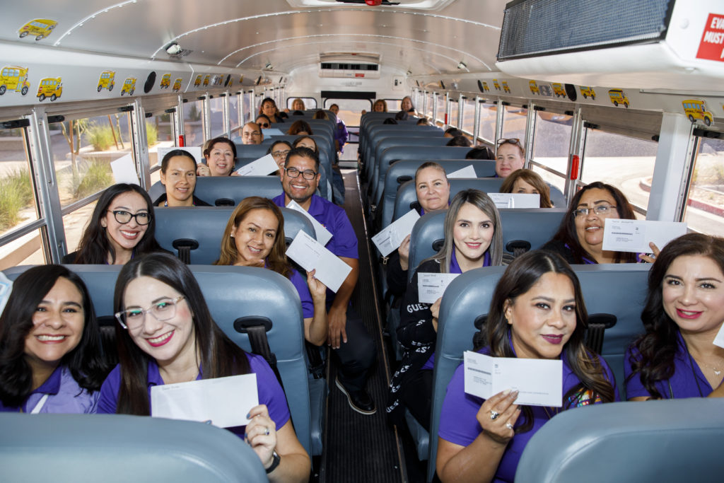 a group of teachers and staff in purple shirts sitting in a school bus hold up white envelopes containing their CREEED gift cards