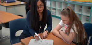 Myla Dayrit, a teacher in a long sleeve blue shirt and black glasses sits next to a student in a white t-shirt and shows a math function on a small dry erase board with a black marker